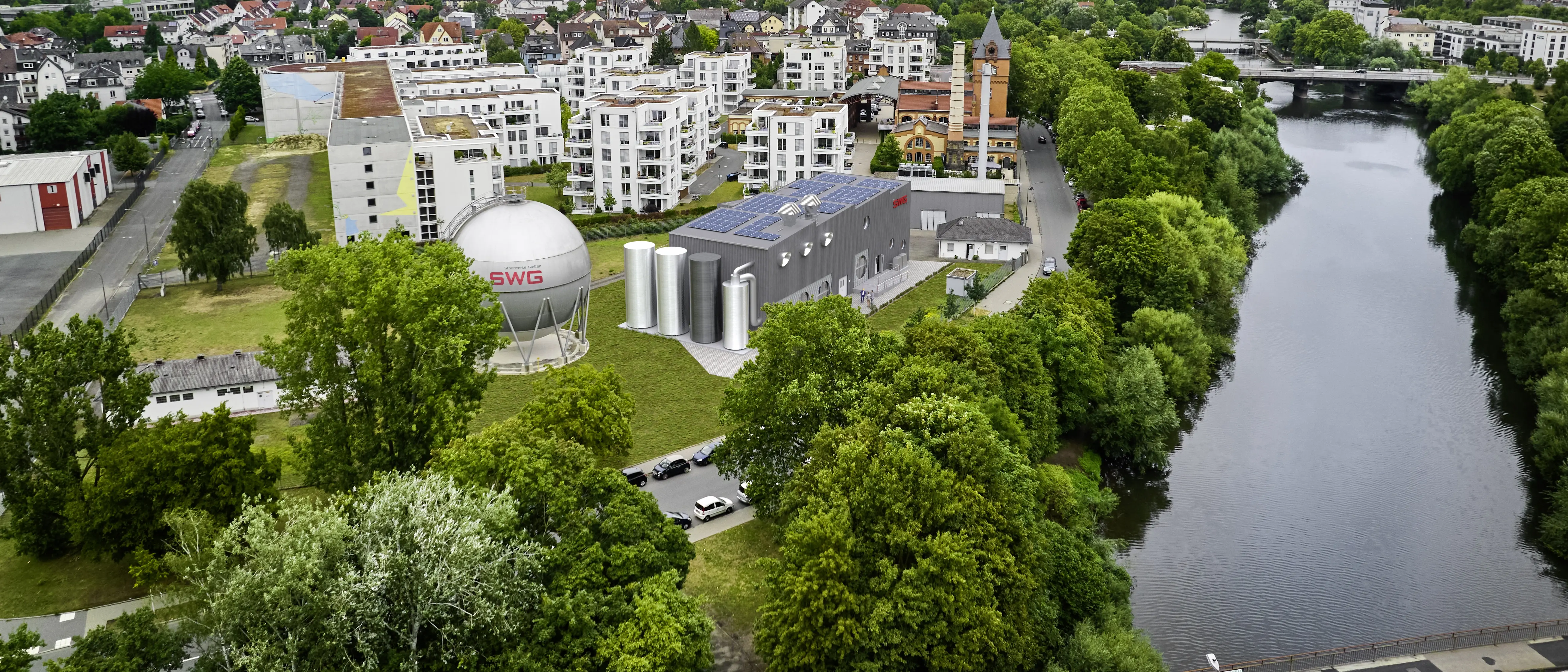 SWG-Flusswasserwärmepumpe Das Bild zeigt eine Luftaufnahme eines Stadtgebiets mit einem zentralen Gebäudeensemble, bestehend aus einem großen kugelförmigen Gasspeicher mit der Aufschrift „SWG“, mehreren zylindrischen Tanks und einem Flachdachgebäude mit Solarpaneelen. Die Gebäude sind umgeben von Wohnhäusern und viel Grün.