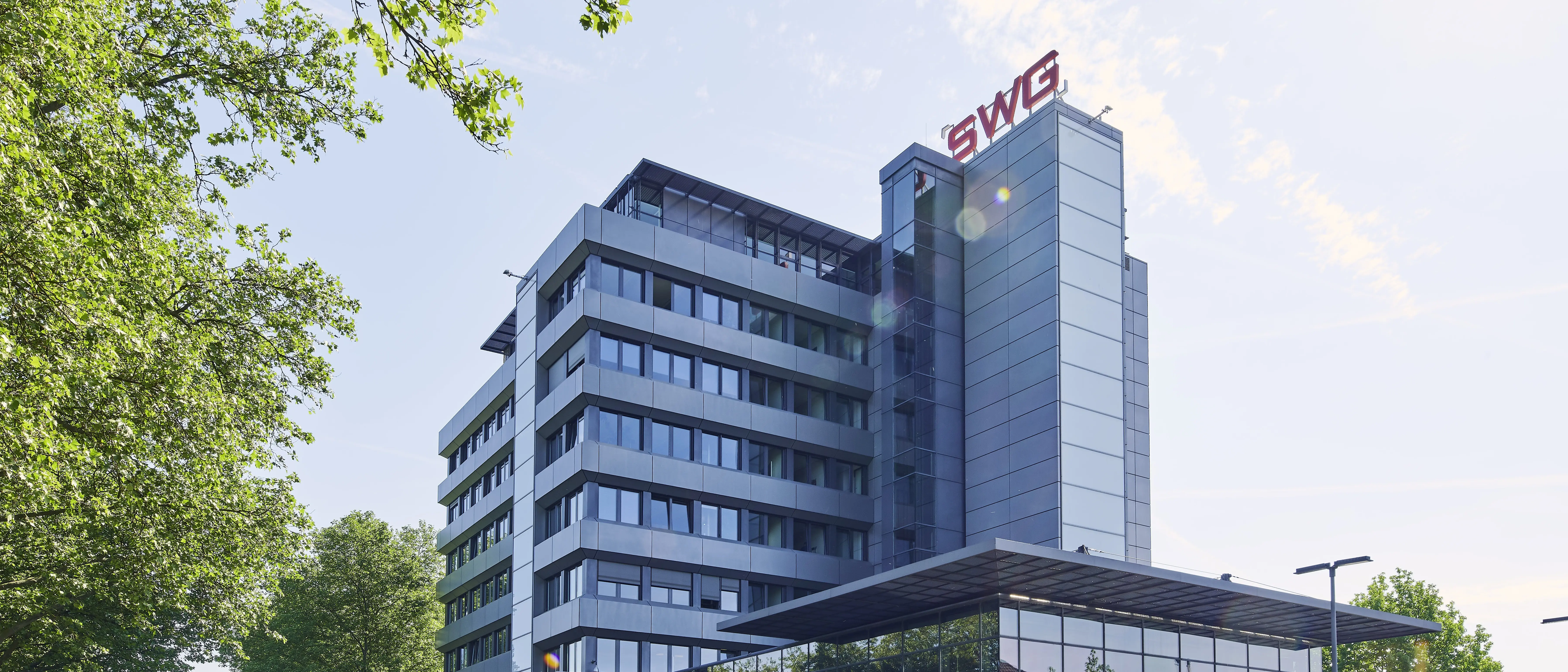 SWG-Hauptgebäude Ein modernes Bürogebäude mit Glas- und Metallfassade, markiert mit dem roten Logo "SWG" auf dem Dach. Umgeben von grünen Bäumen und unter klarem Himmel mit einigen Wolken.